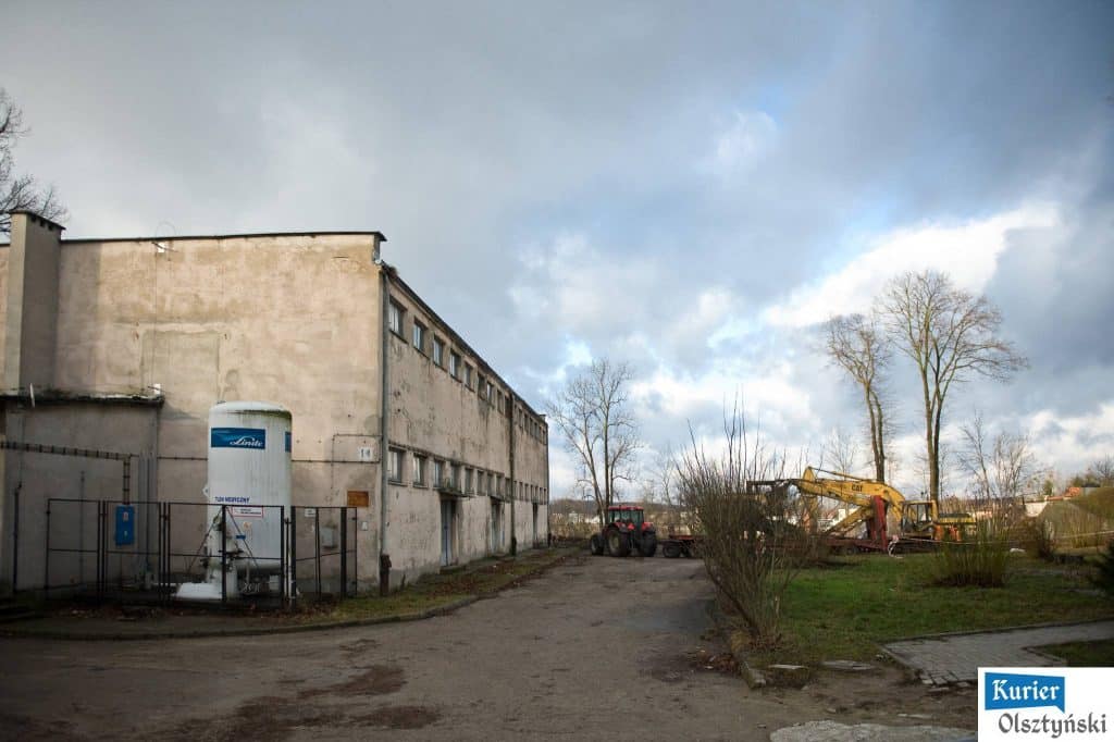 Widok na starą, nieco zniszczoną budowlę szpitalną z pojazdami i drzewami w tle, nadchodząca zmiana.