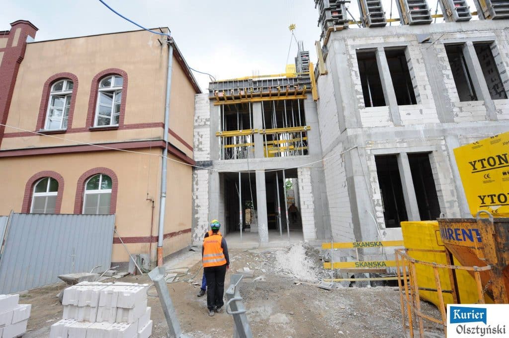 Budowa nowych obiektów szpitalnych na tle poprzednich budynków, widoczne rusztowania i prace na placu budowy.