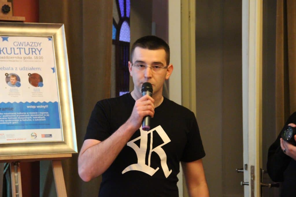 Mężczyzna w czarnym T-shircie z mikrofonem, stojący obok plakatu o kulturalnym wydarzeniu.