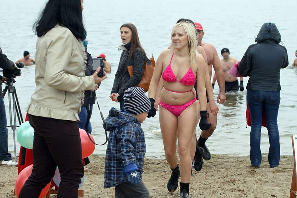 Uczestnicy kąpieli zimowej w Olstynie, w tym kobieta w bikini i dzieci, na plaży przy jeziorze, z kamerami i innymi osobami w tle.
