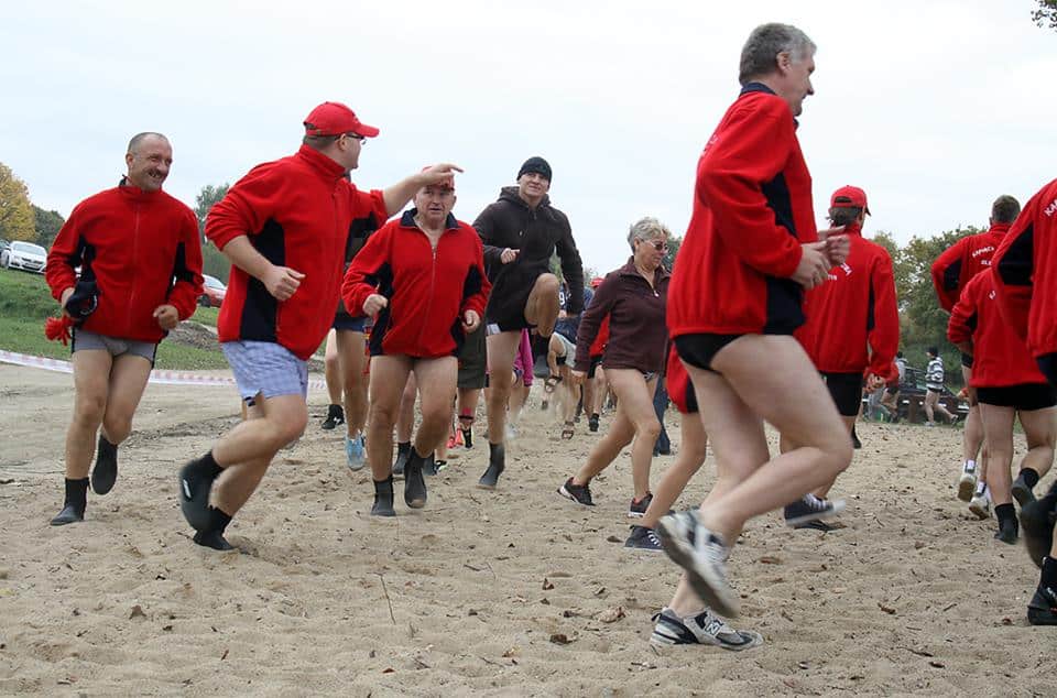 Grupa ludzi w czerwonych kurtkach i bez spodni biegnie po piasku na plaży, przygotowując się do zimowej kąpieli.