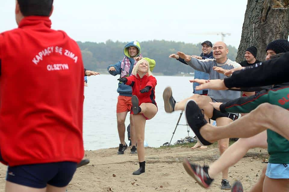 Grupa morsów podczas inauguracji sezonu zimowego na plaży nad jeziorem, uczestnicy ćwiczą i się bawią.