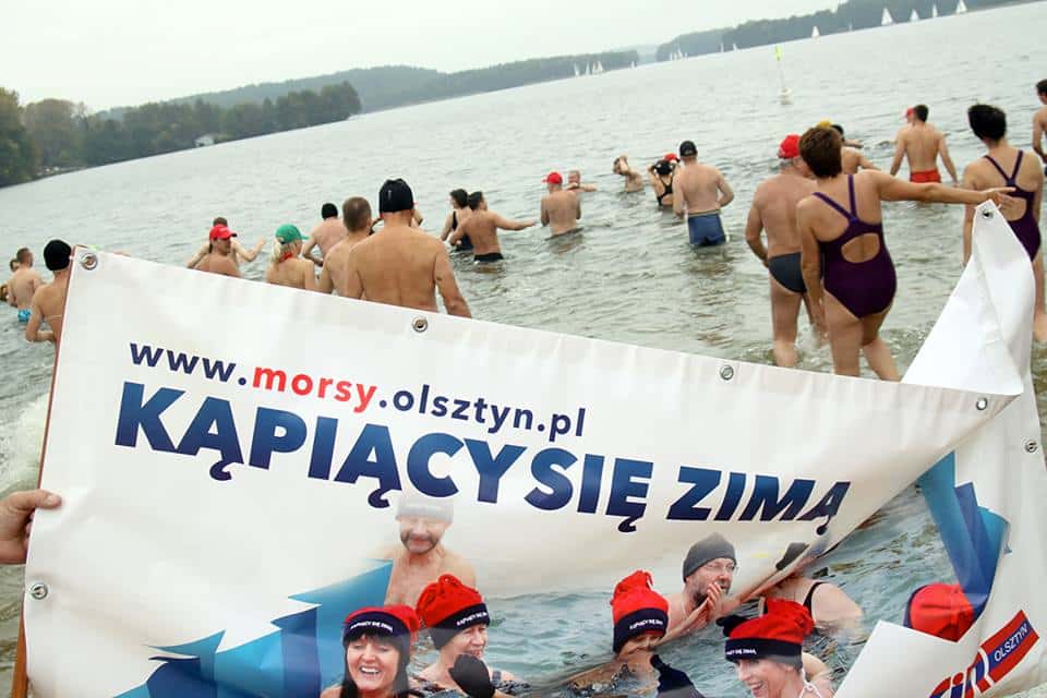 Grupa morsów w zimnej wodzie podczas inauguracji sezonu, trzymająca transparent z napisem „Kąpiąc się zimą”.