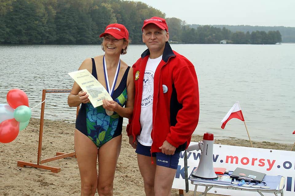 Morsy z Olsztyna podczas inauguracji sezonu kąpielowego nad jeziorem, kobieta z medalem i dokumentem, obok mężczyzna w czerwonej kurtce.