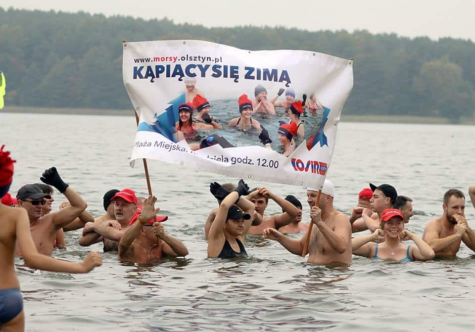 Grupa ludzi w wodzie, trzymająca baner z napisem "Kąpią się zimą" podczas inauguracji sezonu morsowego w Olsztynie.