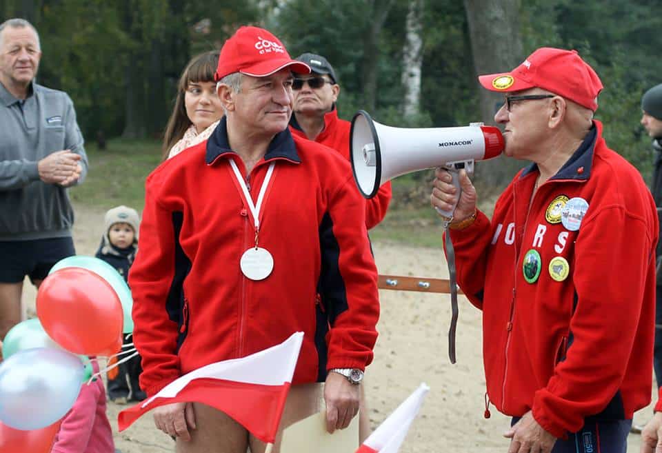 Mężczyzna z megafonem i inny z medalem, ubrani na czerwono, podczas inauguracji sezonu zimowego kąpieliska, z grupą ludzi w tle.