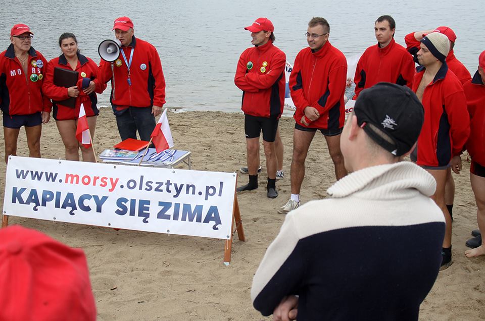 Grupa morsów w czerwonych kurtkach i czapkach na plaży podczas inauguracji sezonu zimowego, w tle woda i nadmorski piasek.