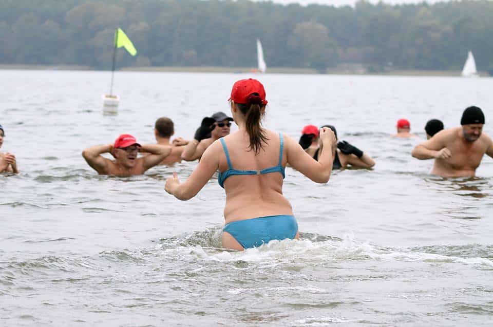 Grupa kąpiących się w jeziorze ludzi, kobieta w bikini i czerwonej czapce w centrum.