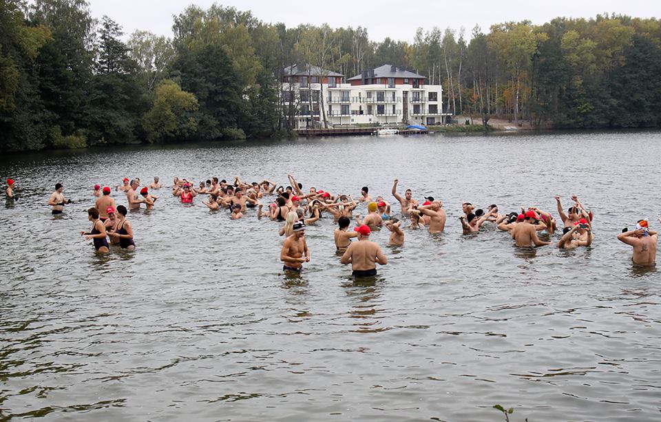 Grupa ludzi kąpie się w jeziorze pod koniec sezonu zimowego, część z nich nosi czapki i stroje kąpielowe.