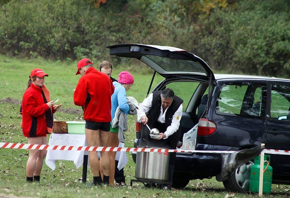 Grupa osób stoi przy samochodzie, jedna osoba nalewa gorącą herbatę z termosu, wokół sznur taśmy ostrzegawczej.