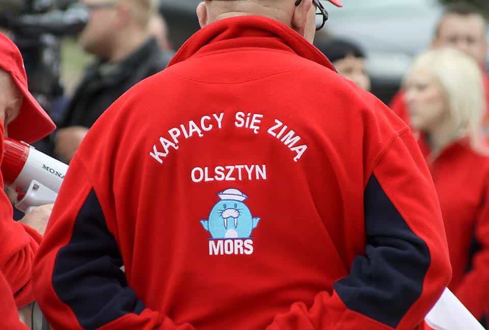 Osoba w czerwonej kurtce z napisem 'Kąpiący się zimą Olsztyn', widoczna od tyłu, w grupie ludzi na zewnątrz.