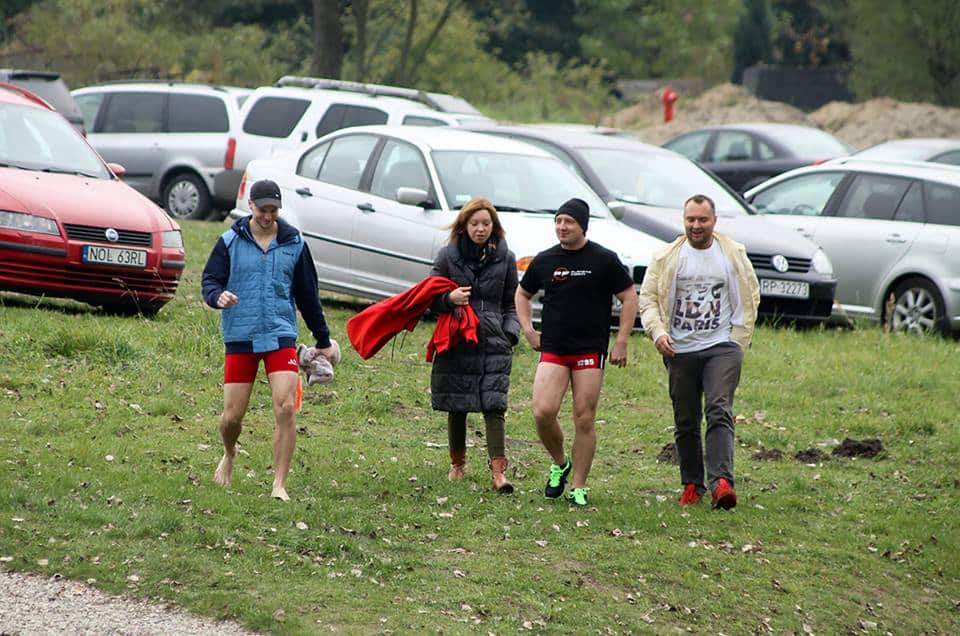 Grupa osób spaceruje po trawie, niektórzy w sportowych szortach, w tle samochody na parkingu.