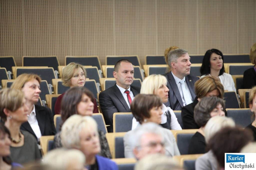 Uczestnicy uroczystości Dnia Edukacji Narodowej siedzą w auli, słuchając wystąpienia. W tle pusta sala z fotelami.
