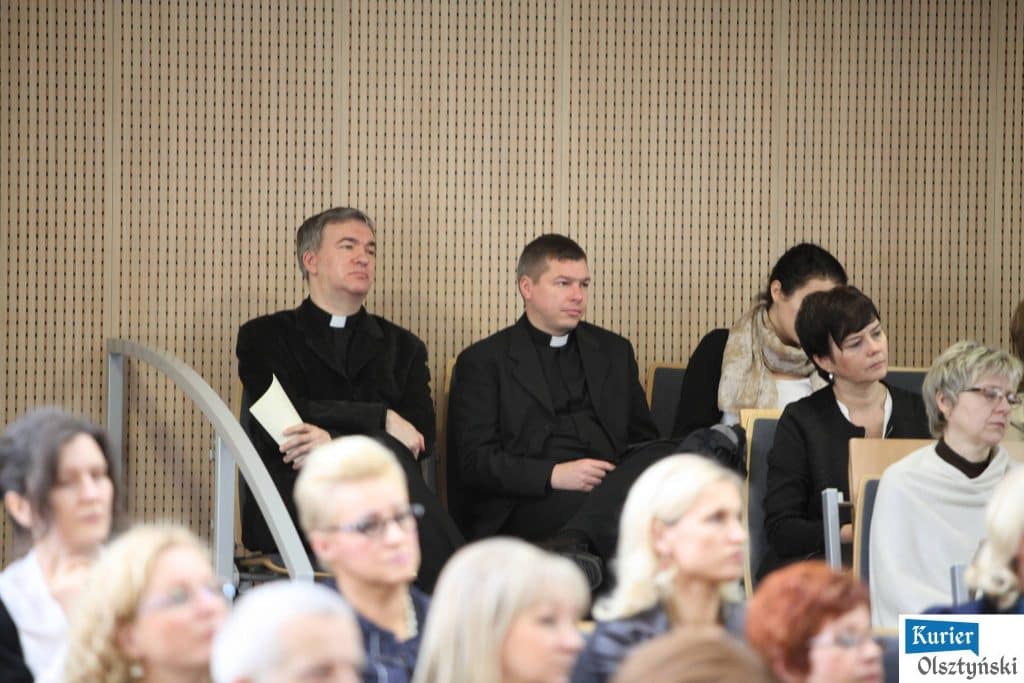 Uczestnicy uroczystości Dnia Edukacji Narodowej, w tym księża i osoby w formalnych strojach, siedzą na widowni w sali konferencyjnej.
