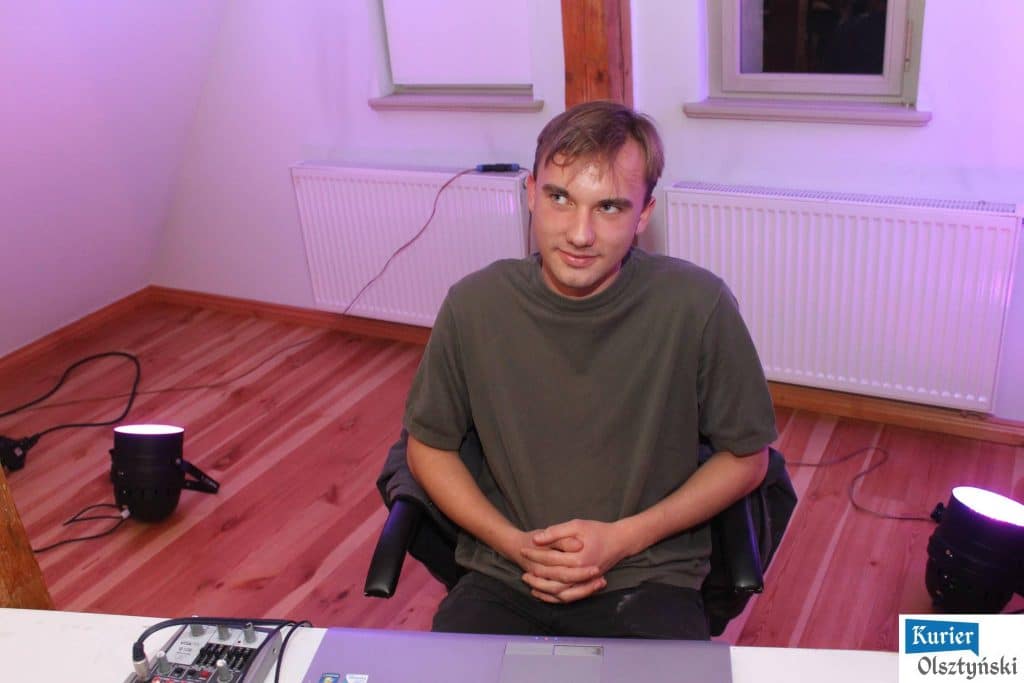 Młody mężczyzna w ciemnej koszulce siedzi przy stole z urządzeniami audio, w tle okno i grzejniki na ścianie.
