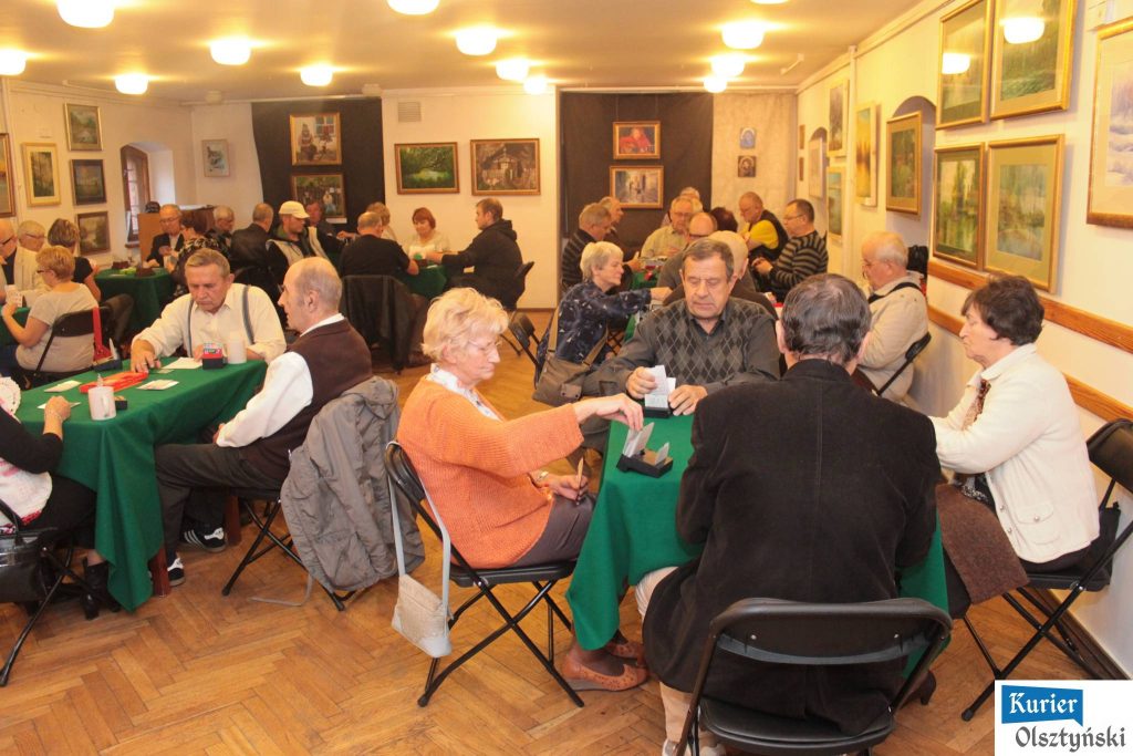 Grupa osób grających w brydża w klubie z obrazami na ścianach.