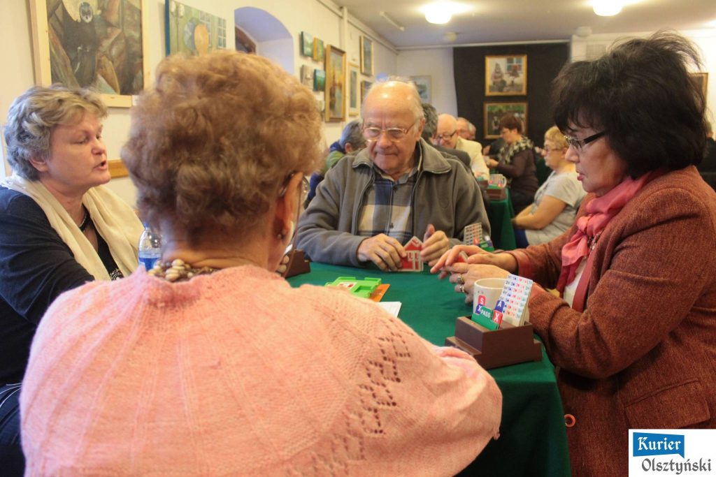 Grupa seniorów gra w brydża przy zielonym stole w klubie kultury, wokół obrazki na ścianach.