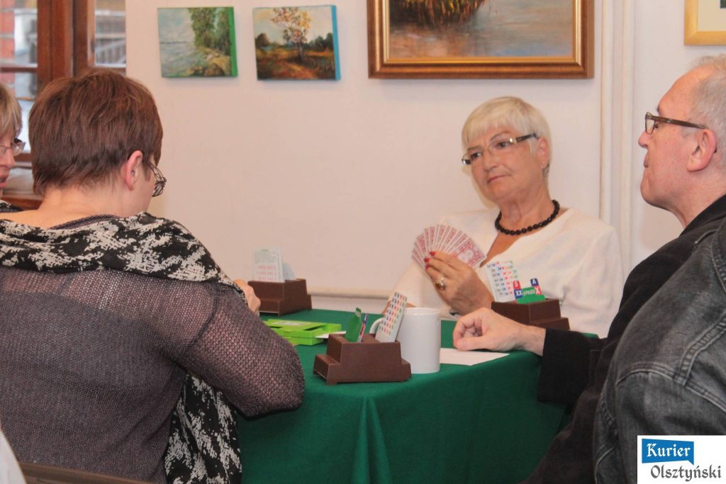 Grupa osób gra w brydża przy stole w pomieszczeniu z obrazami na ścianie.