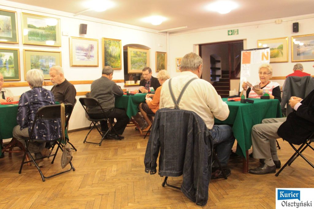 Grupa starszych osób gra w brydża przy stolikach w pomieszczeniu z obrazami na ścianach.