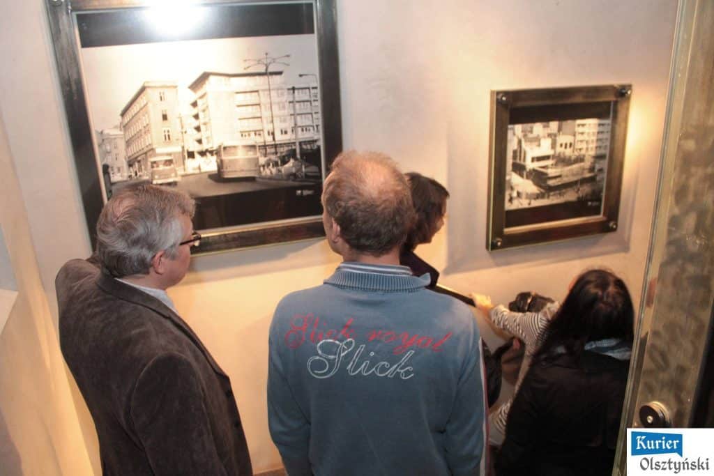 Grupa osób ogląda stare czarno-białe fotografie Olsztyna w galerii na klatce schodowej nowego budynku.