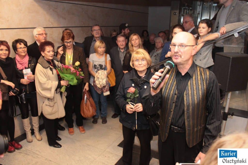 Grupa ludzi na wernisażu zdjęć Zofii Barankiewicz w galerii przy ulicy 1 Maja 6, jedna osoba mówi do mikrofonu.