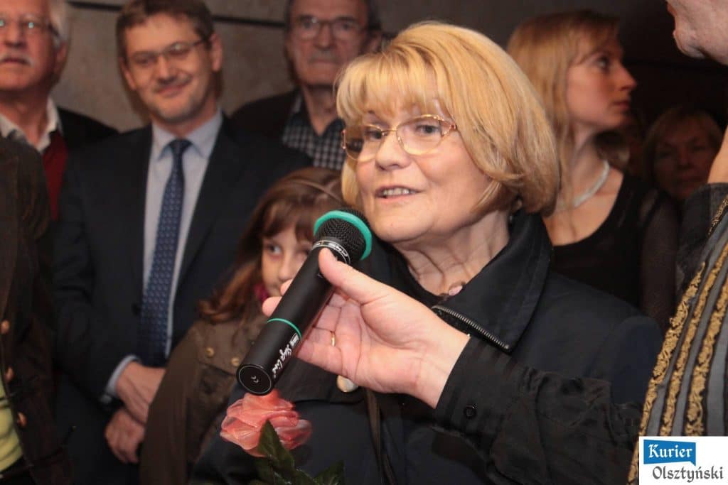 Kobieta z okularami mówi do mikrofonu podczas wernisażu zdjęć dawnego Olsztyna.