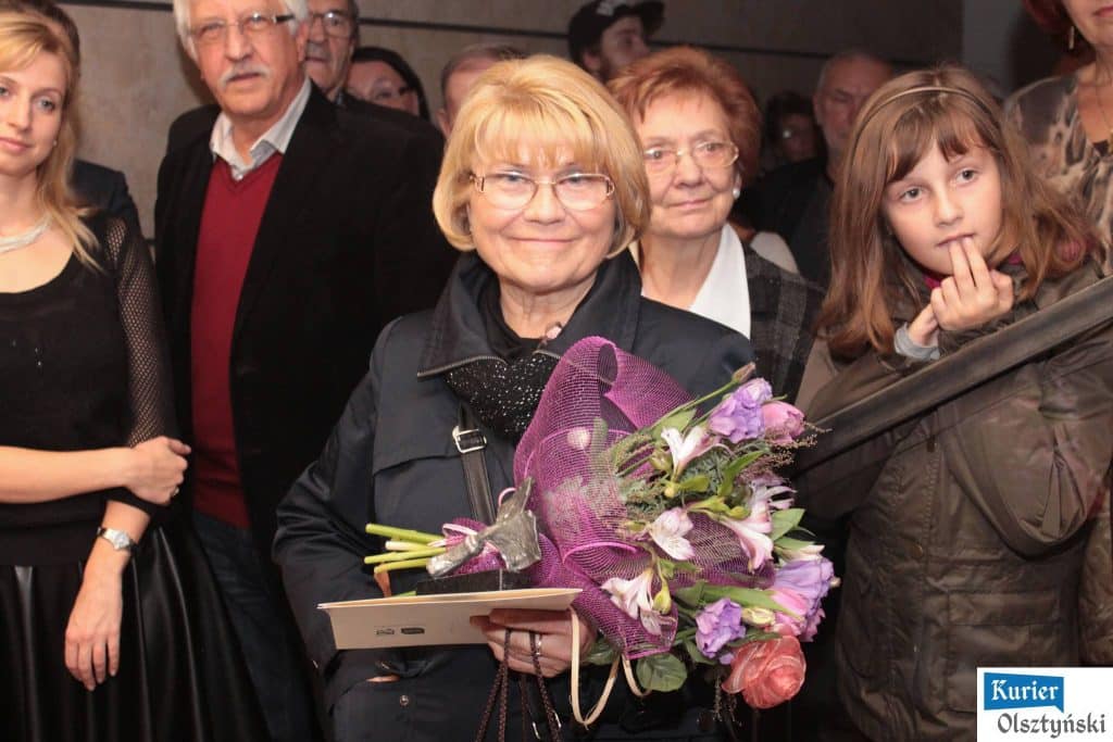 Na zdjęciu grupa ludzi na wernisażu zdjęć Zofii Barankiewicz, w centrum starsza kobieta z kwiatami, wokół jej ludzie.