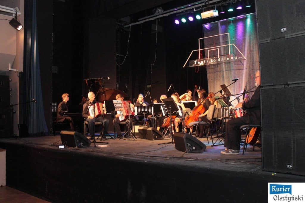 Zespół muzyczny wykonuje utwory na scenie teatralnej, z widownią w tle, podczas koncertu poświęconego pamięci Marka Grechuty.