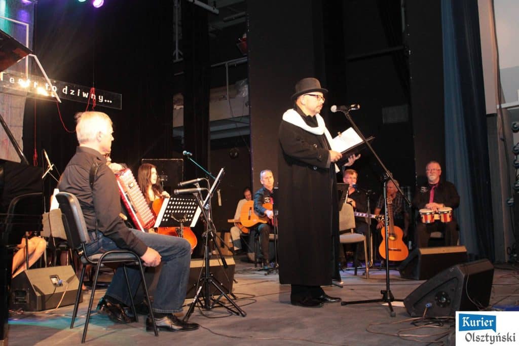 Muzycy na scenie podczas koncertu pamięci Marka Grechuty, z głównym wykonawcą czytającym tekst, wokół instrumenty muzyczne.