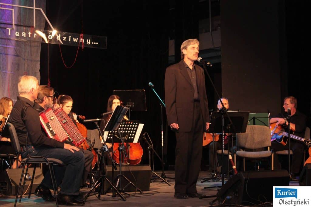 Muzycy i chór na scenie podczas koncertu pamięci o Marku Grechucie, z mężczyzną odczytującym życzenia lub fragment utworu.