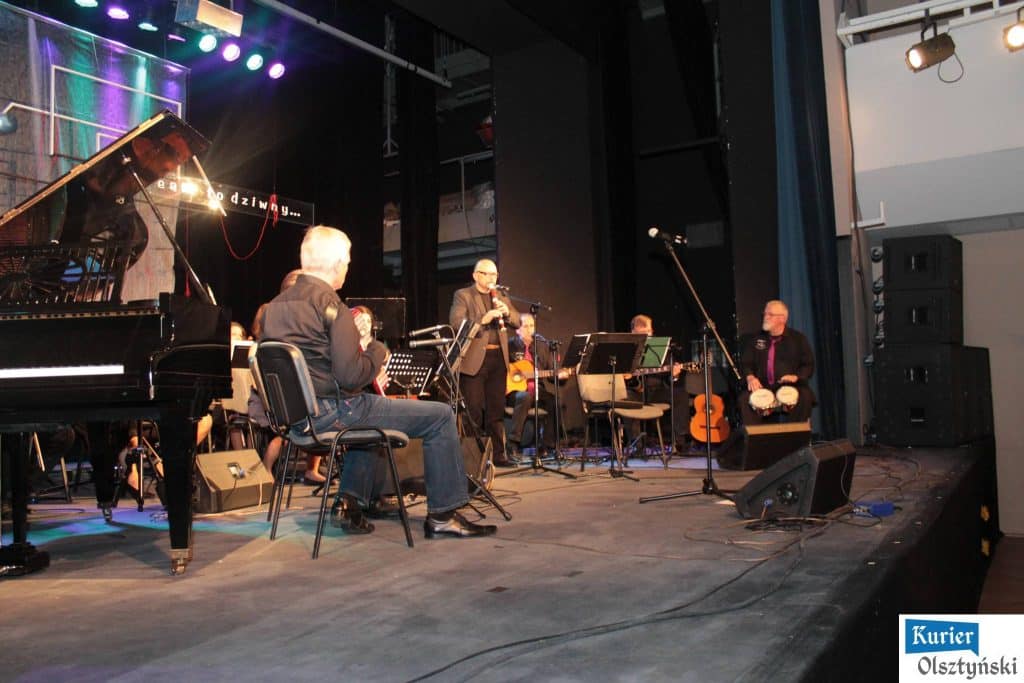 Muzycy na scenie podczas koncertu upamiętniającego Marka Grechutę, z pianinem, gitarami i perkusją, na tle czarnej kurtyny.