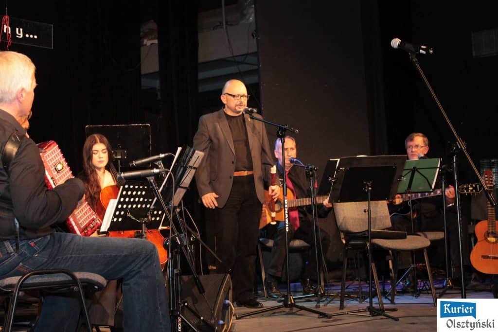 Muzycy podczas koncertu upamiętniającego Marka Grechutę na scenie, z mikrofonem i instrumentami, w ciemnym wnętrzu teatru.