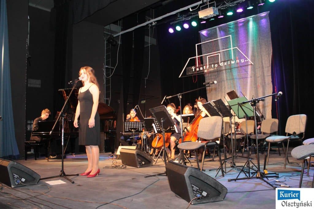 Uczestnicy koncertu na scenie, solistka śpiewa, za nią orkiestra, w tle logo teatru.