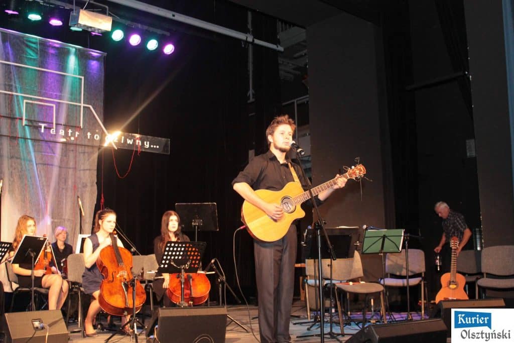 Muzycy na scenie, z solo grający mężczyzna z gitarą akustyczną i orkiestra w tle podczas koncertu upamiętniającego Marka Grechutę.