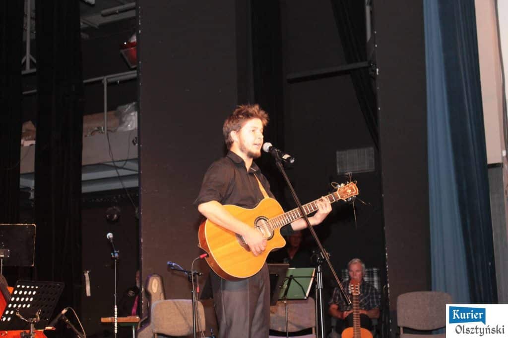 Młody mężczyzna gra na gitarze i śpiewa podczas koncertu w sali teatralnej, za nim scena i inni muzycy.