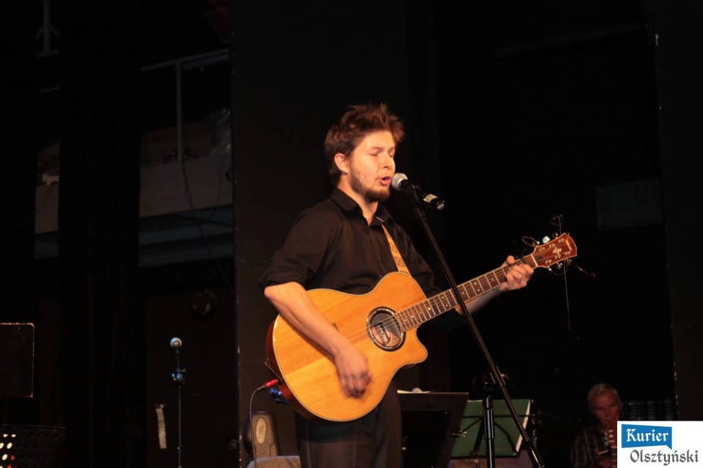 Muzyk śpiewa i gra na gitarze podczas koncertu w teatrze, w tle ciemne ściany i inny muzyk na tle.