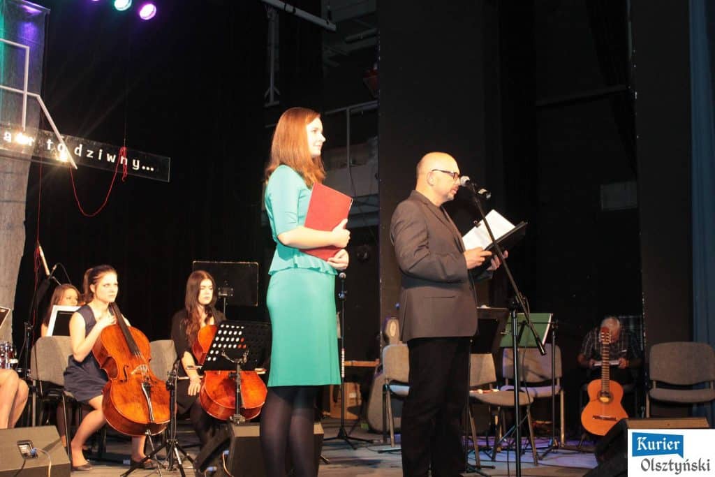 Na scenie muzycy i prelegenci podczas koncertu ku pamięci Marka Grechuty, w tle orkiestra na scenie.