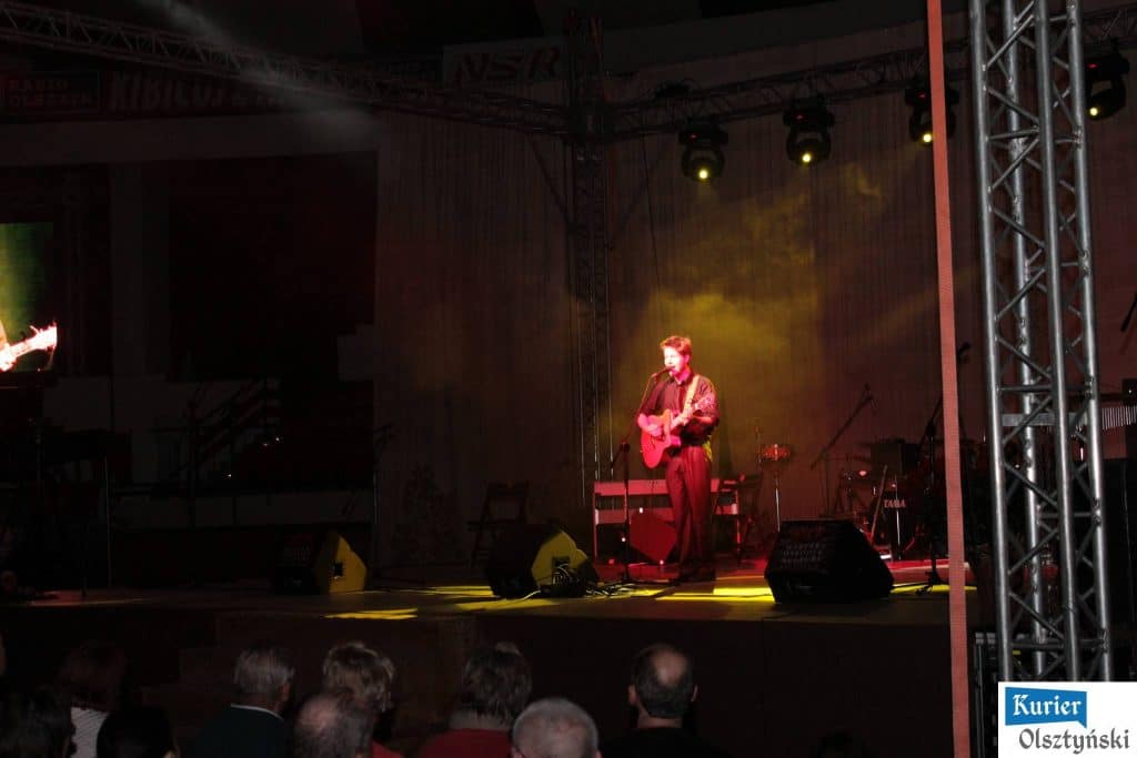 Mężczyzna gra na gitarze podczas koncertu z widownią i oświetleniem na tle sceny.