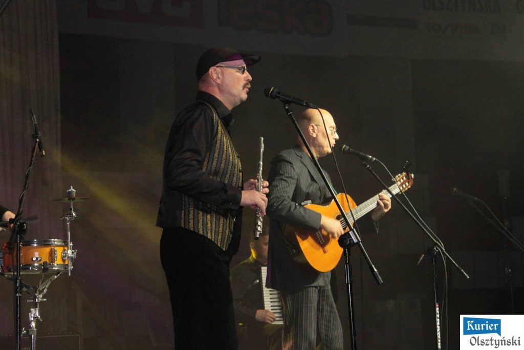 Muzycy na scenie podczas koncertu z gitarami i instrumentem perkusyjnym, w strojach formalnych.