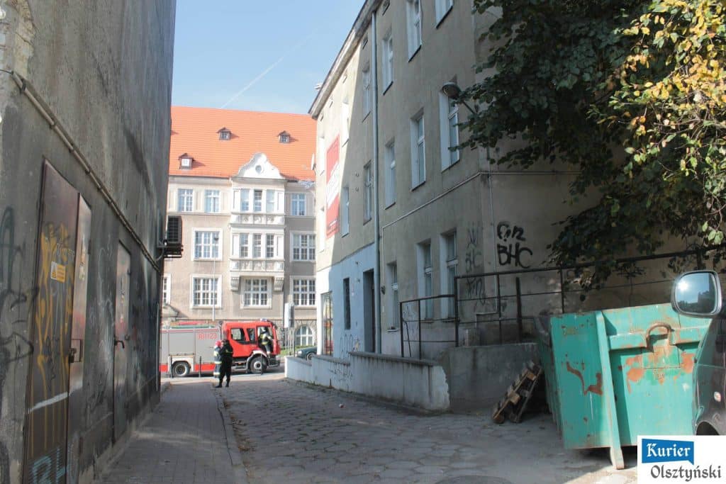 Wąska ulica w centrum Olsztyna z wozami strażackimi i ratownikami podczas akcji ratunkowej.