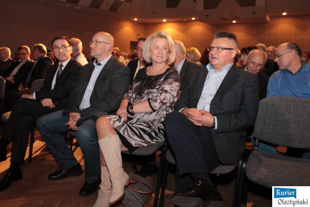 Grupa ludzi siedząca na sali podczas wydarzenia, w centrum kobieta z blond włosami, wokół niej mężczyźni w garniturach.