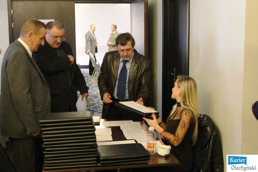 Grupa mężczyzn rozmawia przy rejestracji na konferencji, stojąc przy stoliku z dokumentami i teczkami.