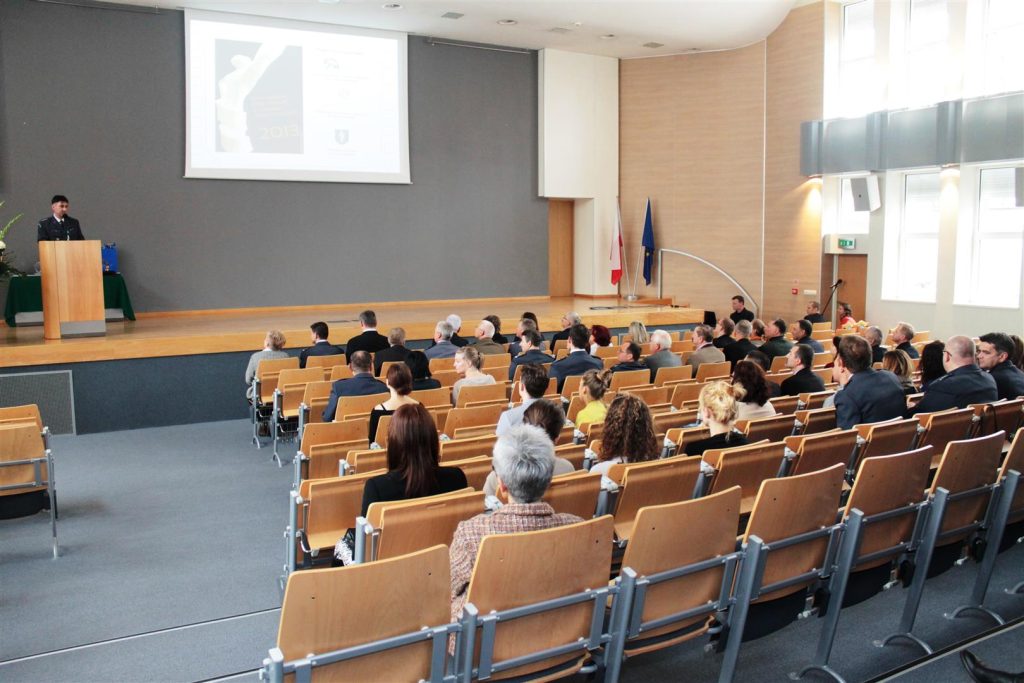 Sala konferencyjna z widokiem na dużą prezentację na ekranie, wypełnioną głównie osobami w formalnych strojach.