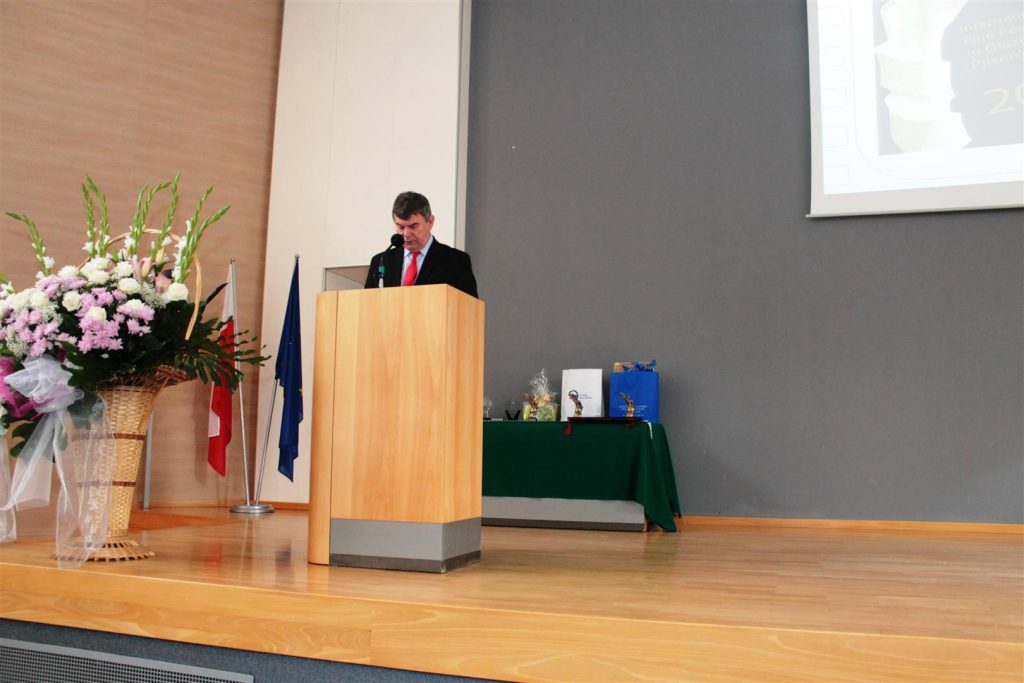 Mężczyzna przemawia z podium podczas gali w urzędzie wojewódzkim, obok kwiaty i flagi.