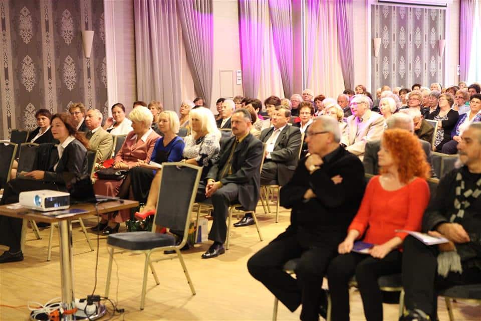 Uczestnicy uroczystości jubileuszowej w auli, siedzą na krzesłach, słuchają wykładu lub wystąpienia.