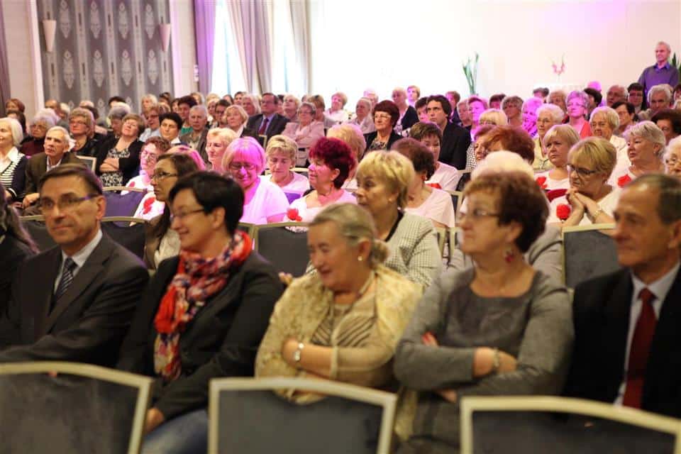 Słuchacze Akademii Trzeciego Wieku podczas uroczystości w sali konferencyjnej, część z nich z uśmiechami, w tle kolorowe światła.