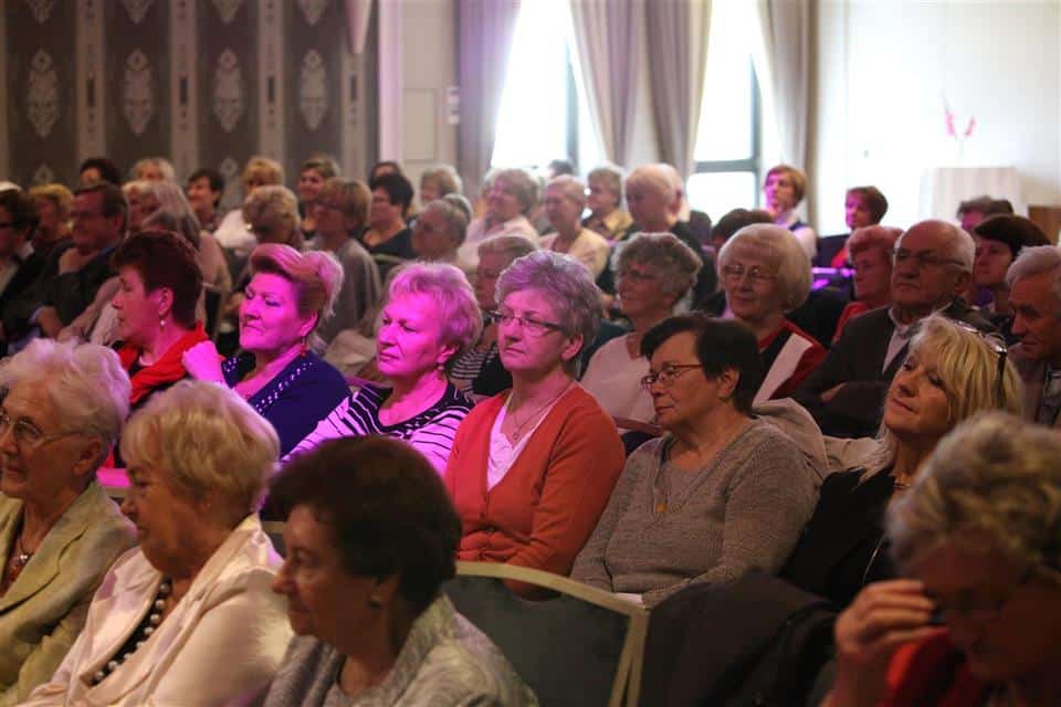 Grupa seniorów podczas wydarzenia w sali konferencyjnej, siedząca na widowni, słuchając z uwagą.
