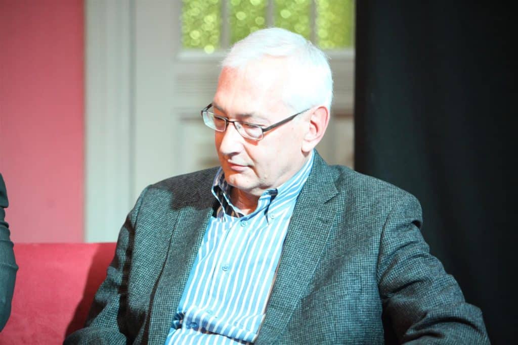 Mężczyzna z krótkimi siwymi włosami noszący okulary, ubrany w marynarkę i koszulę, siedzi w pomieszczeniu.
