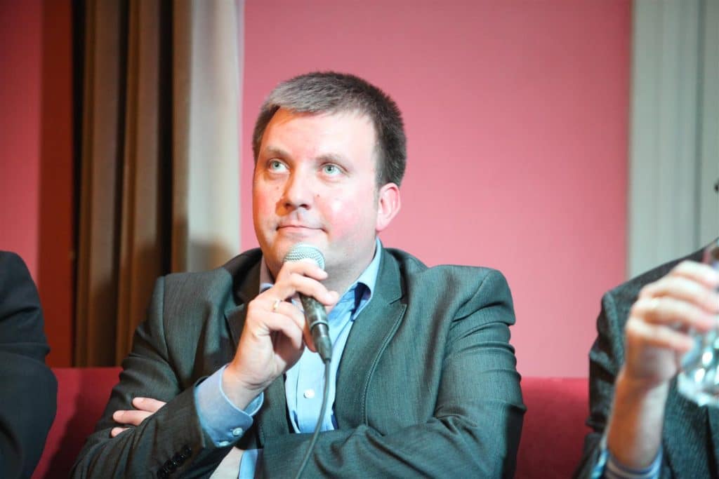 Mężczyzna z krótkimi ciemnymi włosami, ubrany w garnitur, trzyma mikrofon podczas dyskusji na wydarzeniu.