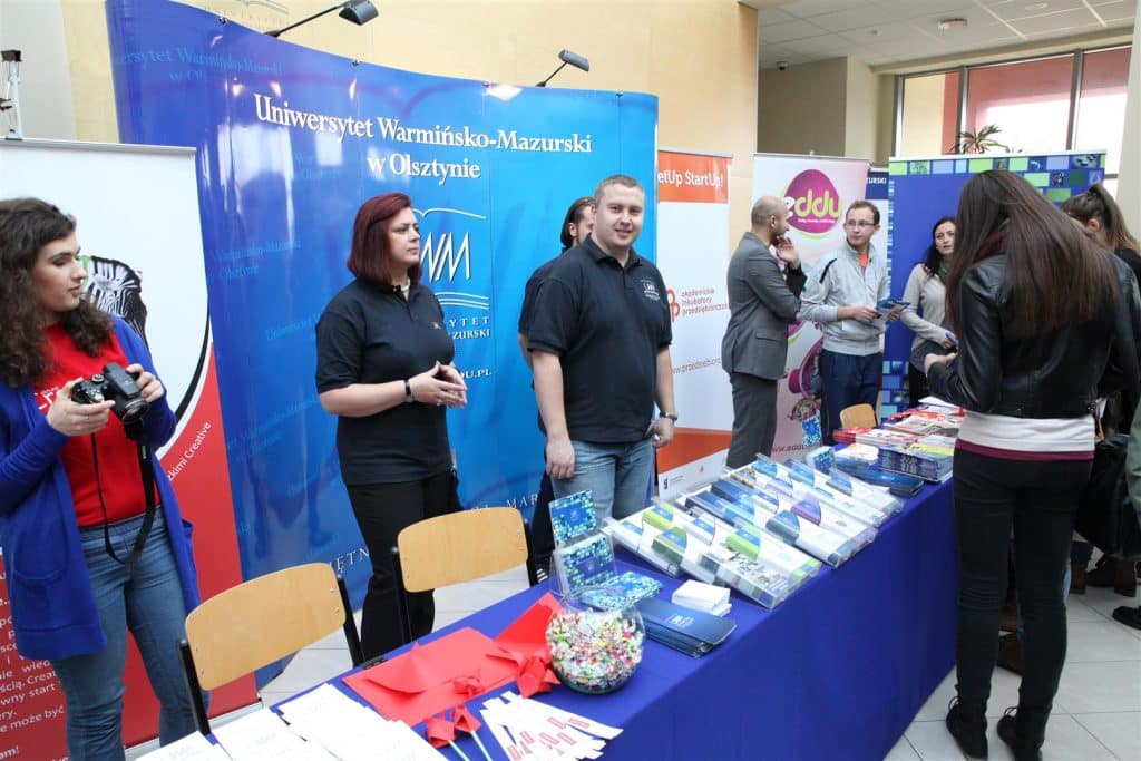 Stoisko z ofertami edukacyjnymi i informacyjnymi na targach w Kortowie, uczestnicy rozmawiają i zwiedzają.